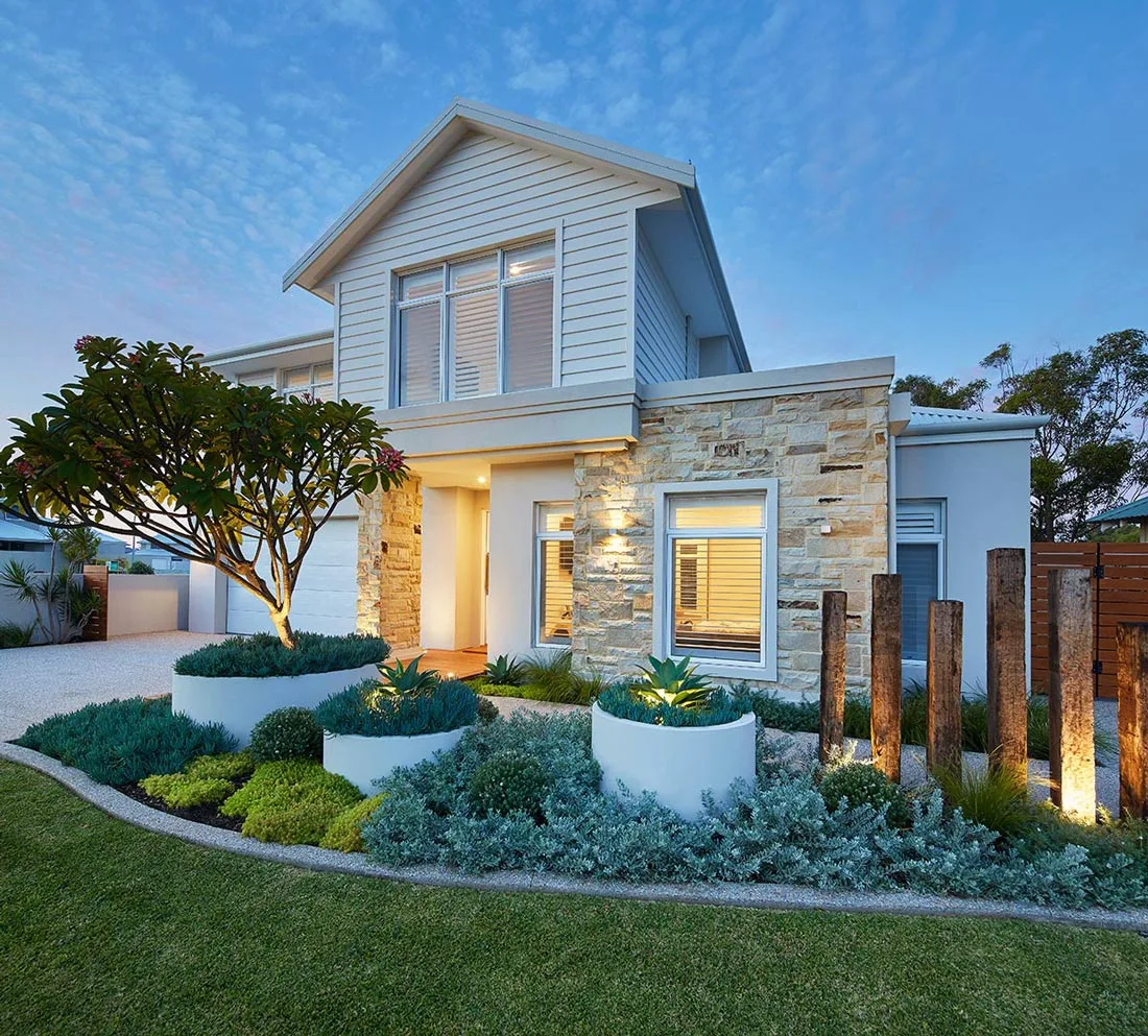 Two-storey home with stone feature wall, timber posts and garden beds lit at dusk